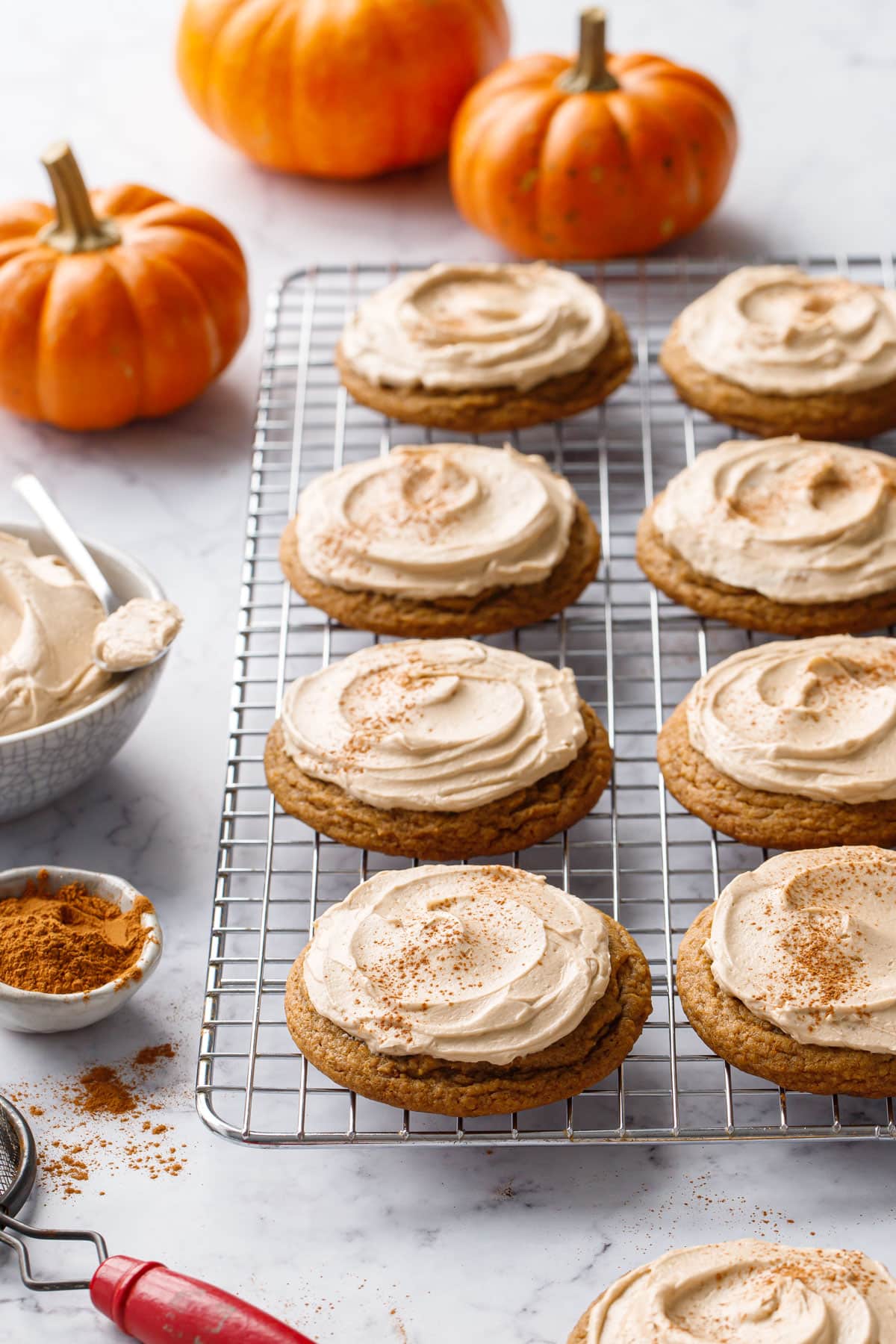 Wire baking rack with rows of frosted Caramel Pumpkin Cookies dusted with cinnamon, decorative orange pumpkins and bowl of frosting in the background.