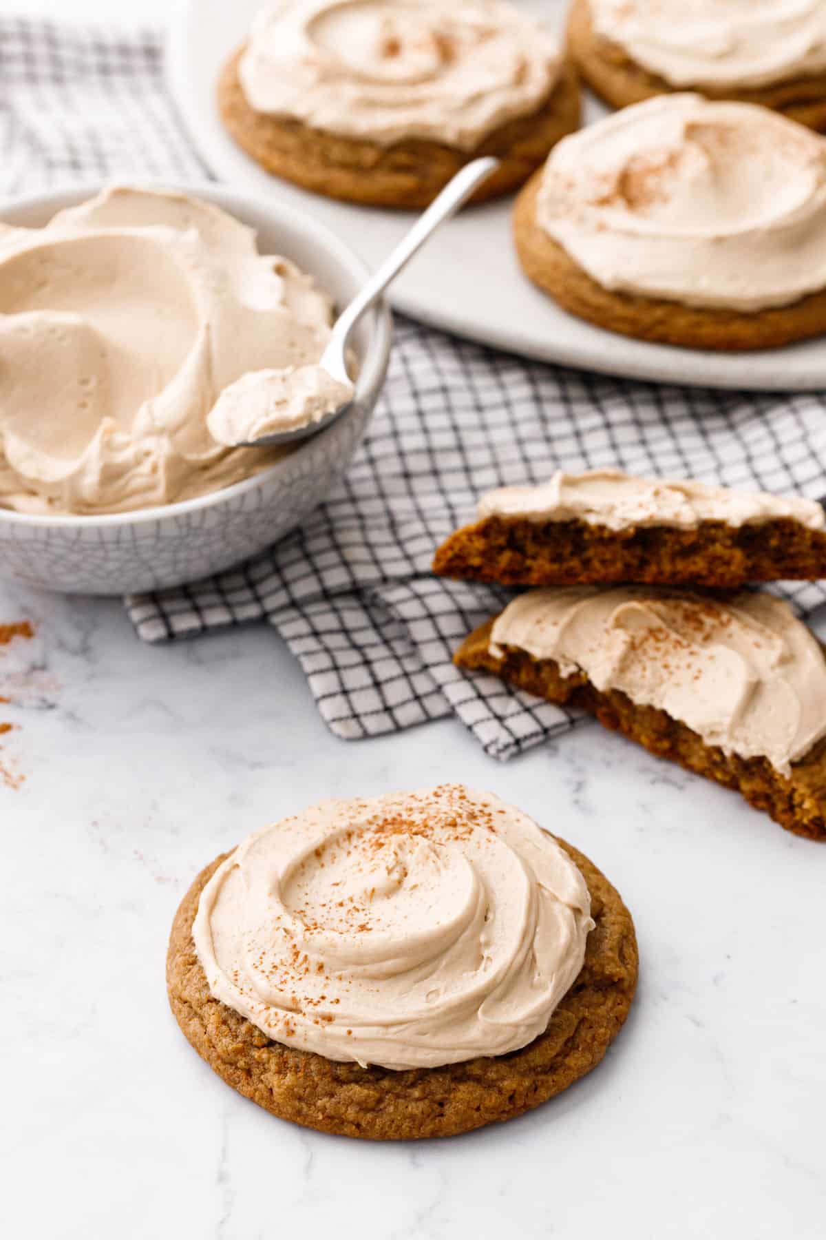 Caramel Pumpkin Cookies, one broken in half, a few on a plate in the background and a bowl of caramel frosting with a spoon.