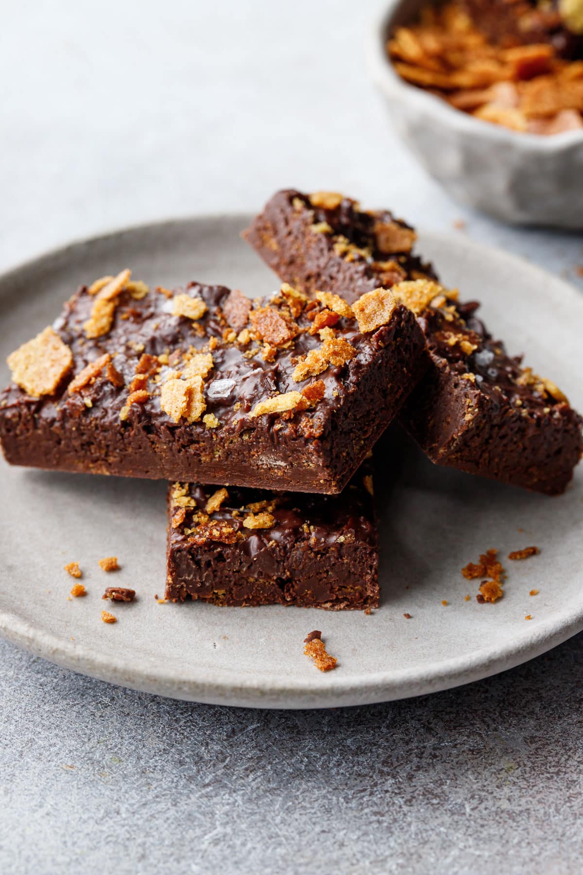 Closeup of three rectangular bars of homemade Nutella Chocolate Crunch bars on a gray ceramic plate, bowl of feuilletine out of focus in the background.