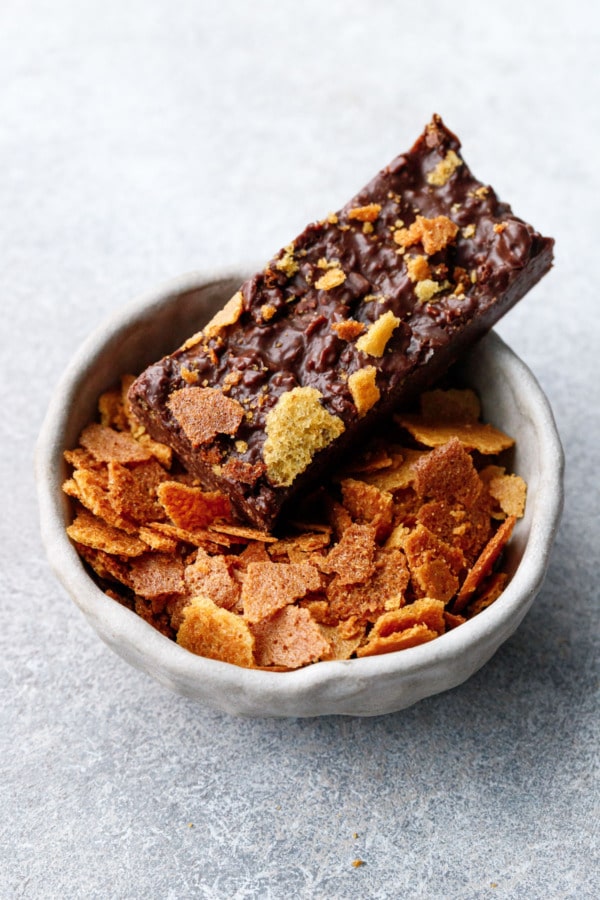 One rectangular Nutella Chocolate Crunch candy bar sitting on the edge of a small bowl of feuilletine flakes.