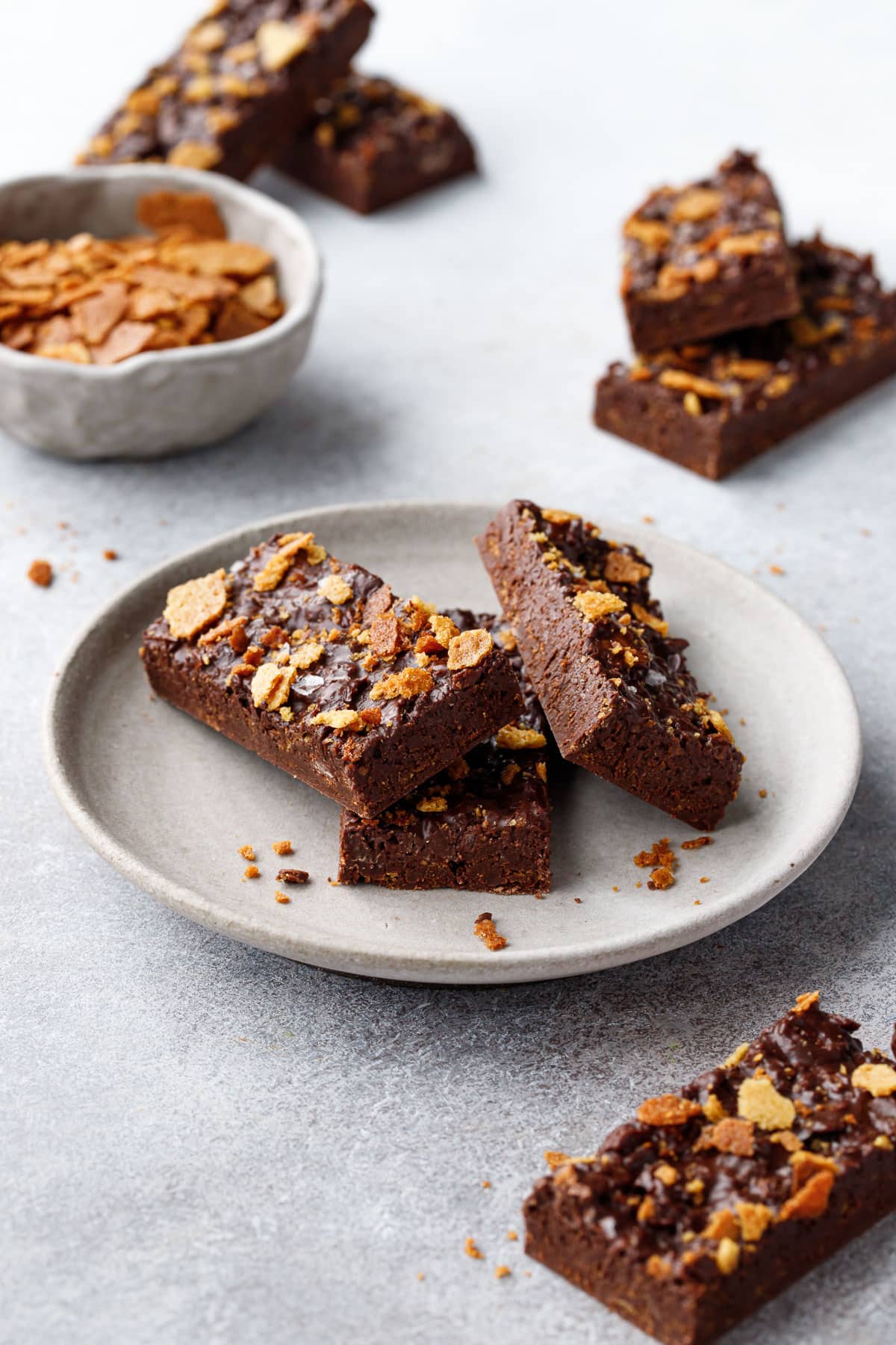 Rectangular cut bars of Nutella Chocolate Crunch  scattered on a gray background, some piled on a plate and some on the side, with a small bowl of feuilletine in the background.