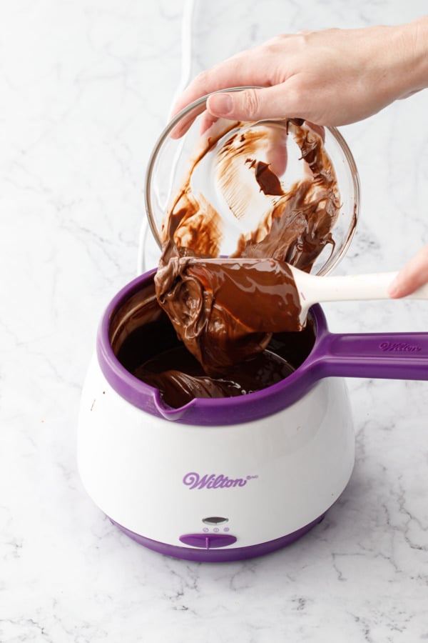 Spatula scraping Nutella from a glass bowl into pot of melted chocolate.