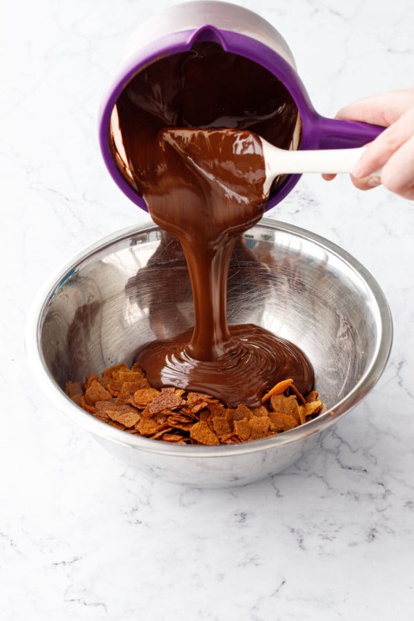 Pouring melted chocolate and Nutella mixture into a stainless steel bowl with feuilletine flakes.