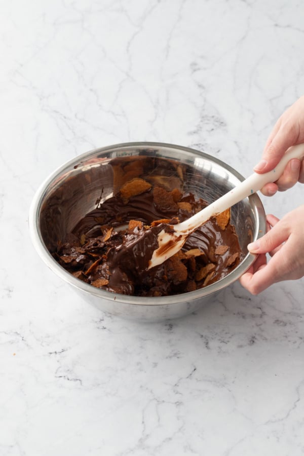 Spatula folding melted chocolate mixture into feuilletine flakes until evenly coated.
