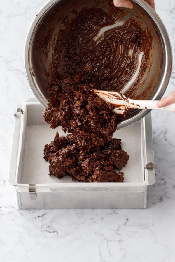 Pouring feuilletine and melted chocolate mixture from stainless mixing bowl into a parchment lined square baking pan.