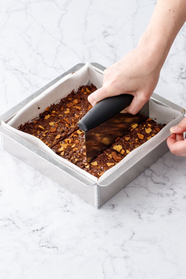 Bench scraper cutting evenly sized bars in a square baking pan before the chocolate has fully set.