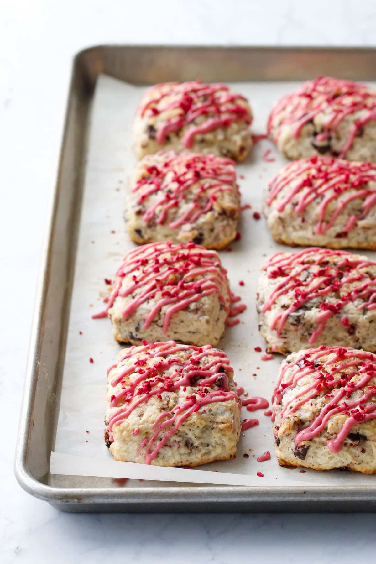 Rows of Dark Chocolate Raspberry Cream Scones on a parchment-lined cookie sheet drizzled with pink raspberry glaze.