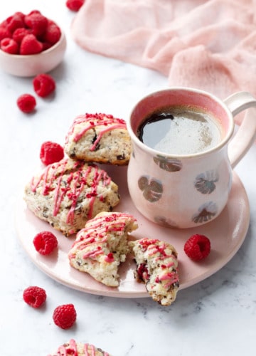 Dark Chocolate Raspberry Cream Scones on a pink serving plate with fresh raspberries and a ceramic mug of coffee, pink napkin and bowl of raspberries in the background.