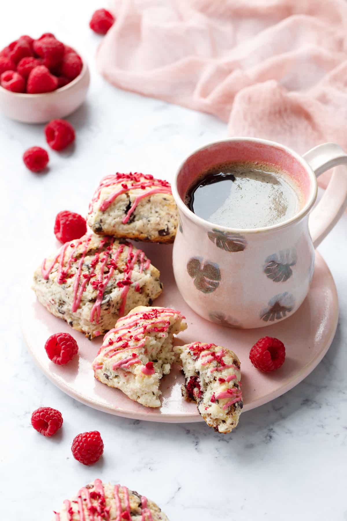 Dark Chocolate Raspberry Cream Scones on a pink serving plate with fresh raspberries and a ceramic mug of coffee, pink napkin and bowl of raspberries in the background.