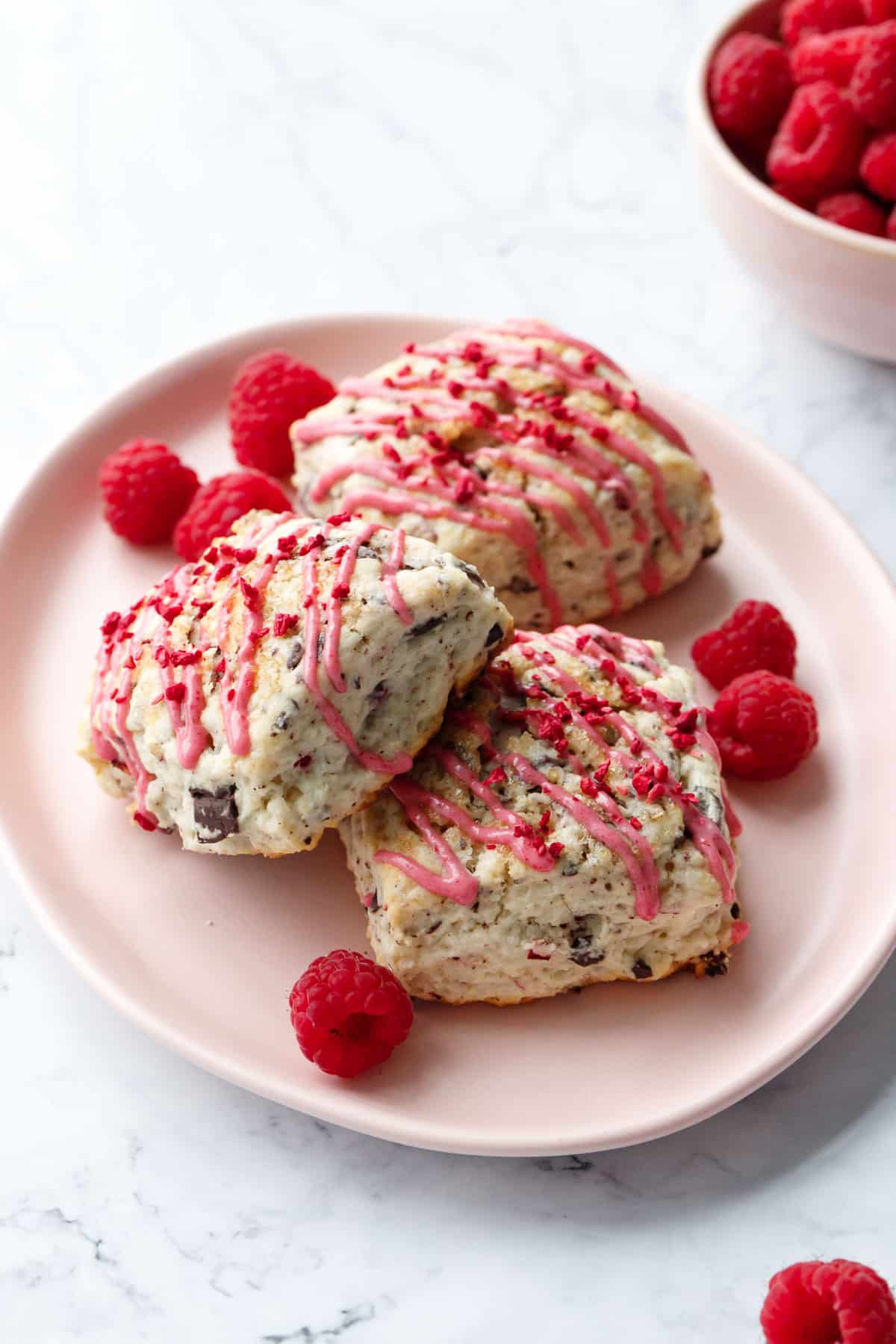 Three Dark Chocolate Raspberry Cream Scones on a pink plate with fresh raspberries on the plate and in a bowl in the background.