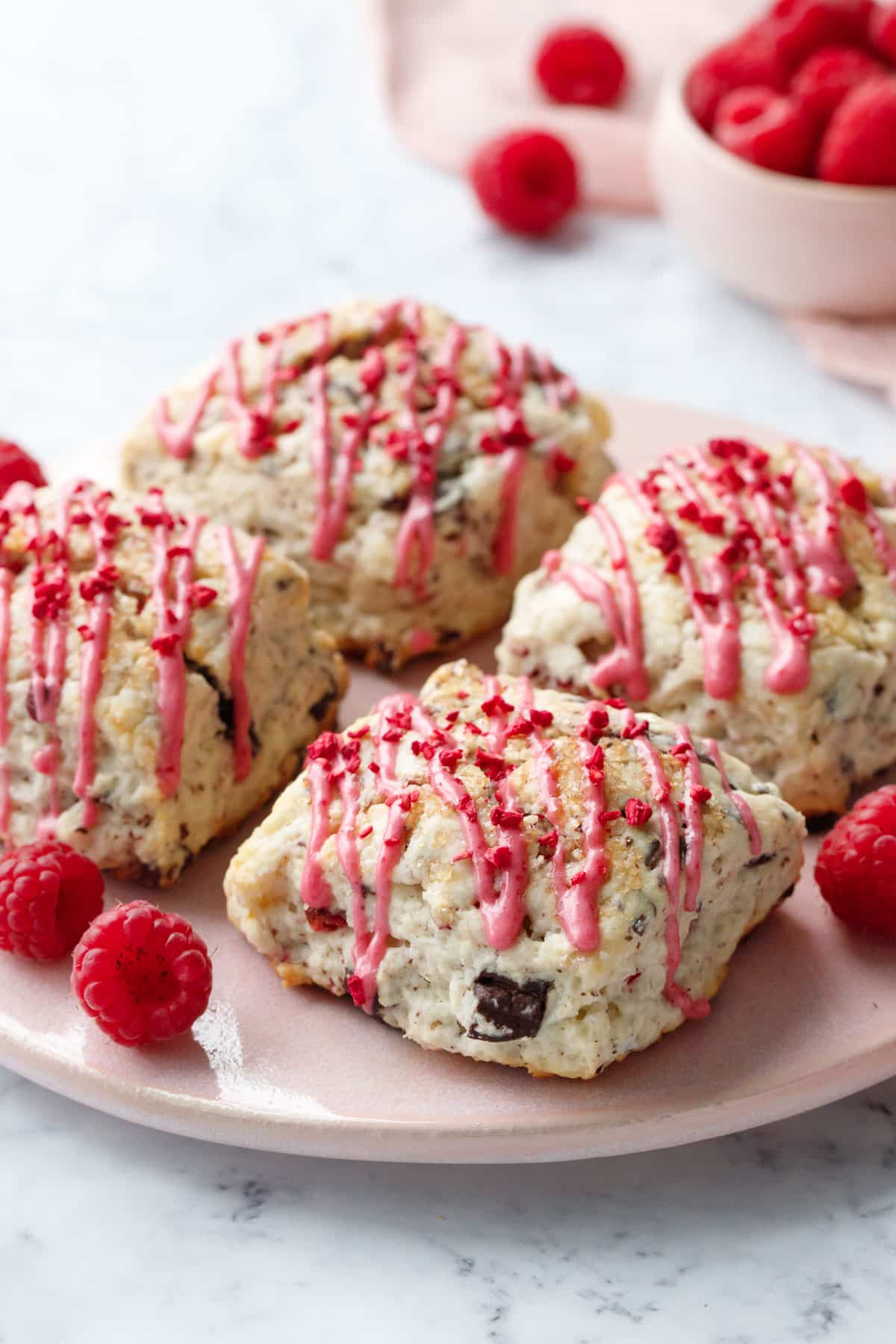 Four Dark Chocolate Raspberry Cream Scones on a pink serving plate, drizzled with pink raspberry glaze and a few fresh raspberries on the side.