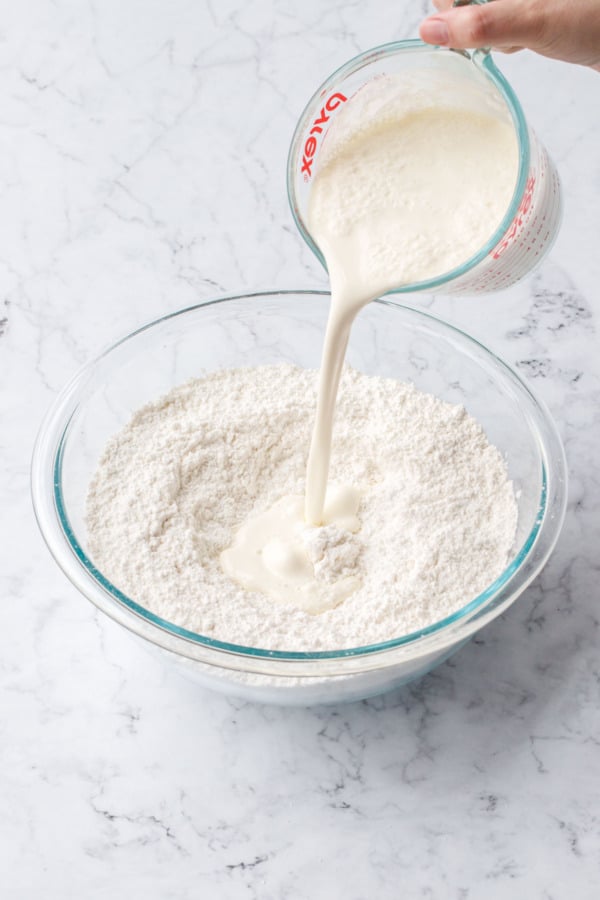 Pouring 2 cups of heavy cream from a glass measuring cup into a mixing bowl with dry ingredients in it.