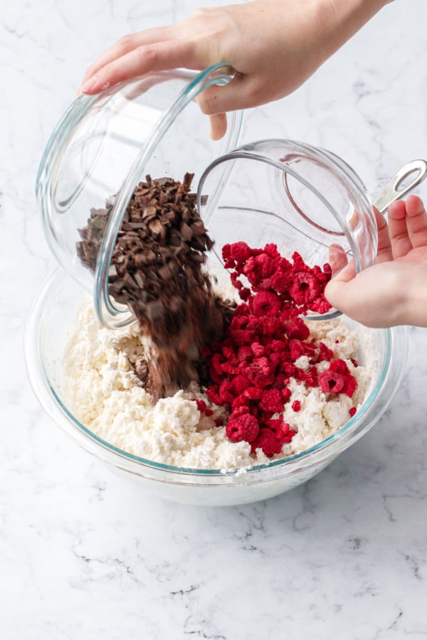 Pouring chopped dark chocolate and crushed freeze-dried raspberries into mixing bowl with partially mixed scone dough.