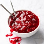 White bowl filled with a chunky cranberry sauce and two spoons on a white marble background.