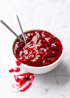 White bowl filled with a chunky cranberry sauce and two spoons on a white marble background.