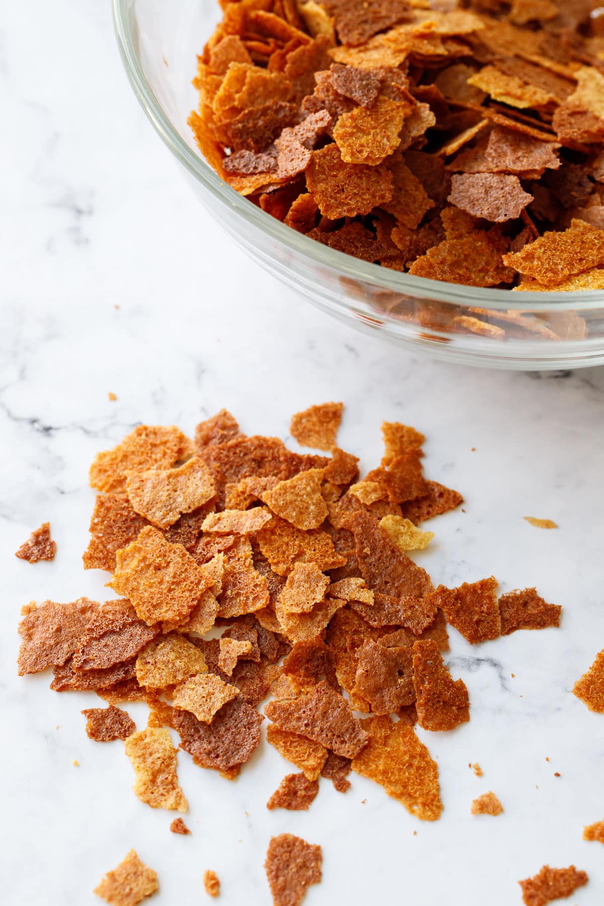 Homemade Feuilletine flakes in a pile on a marble surface with a glass bowl filled with more feuilletine flakes in the background.