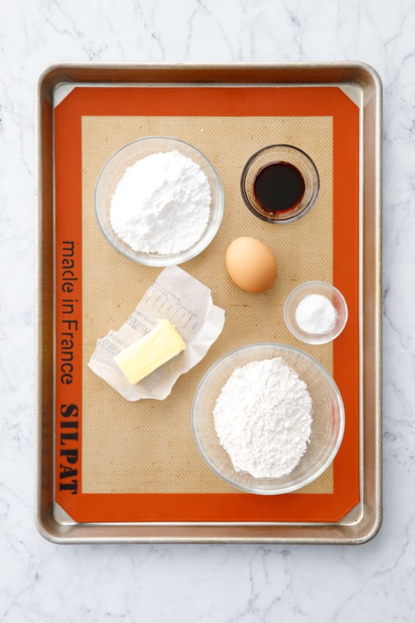 Ingredients to make Homemade Feuilletine (butter, powdered sugar, flour, egg, baking powder, and molasses) set out on a baking sheet.