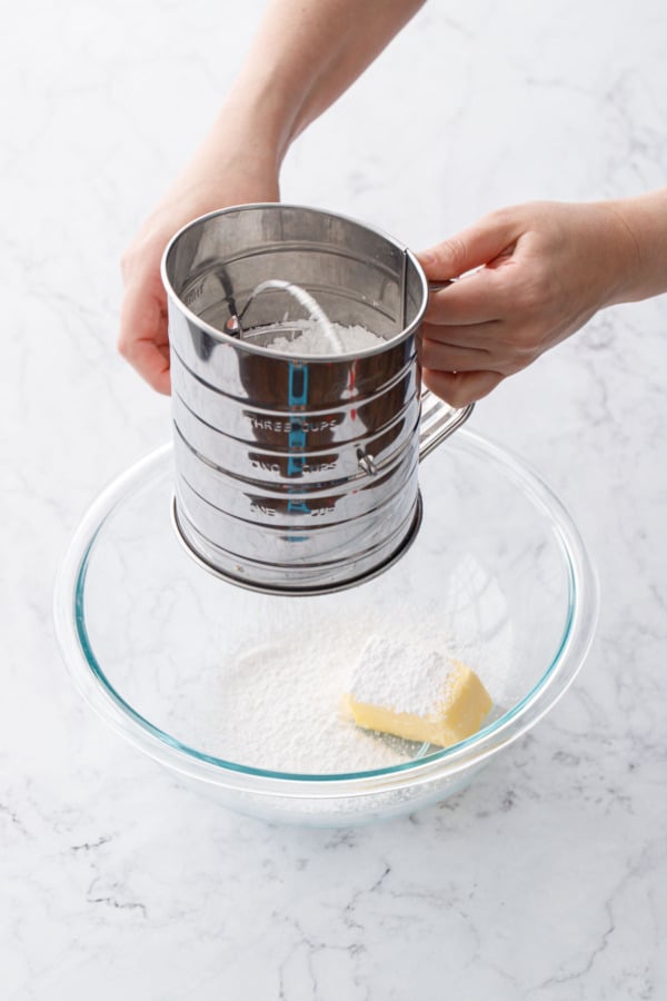 Sifting powdered sugar into a glass bowl with softened butter.