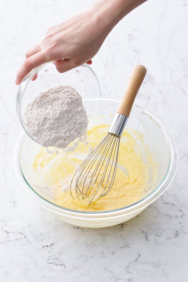 Pouring dry ingredients into bowl with sugar, butter, and egg mixture and a whisk.