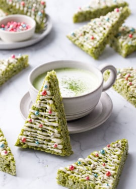 Matcha Rice Crispy Treats cut into triangles and decorated like Christmas trees, one leaning up against a mug of matcha on a marble background.