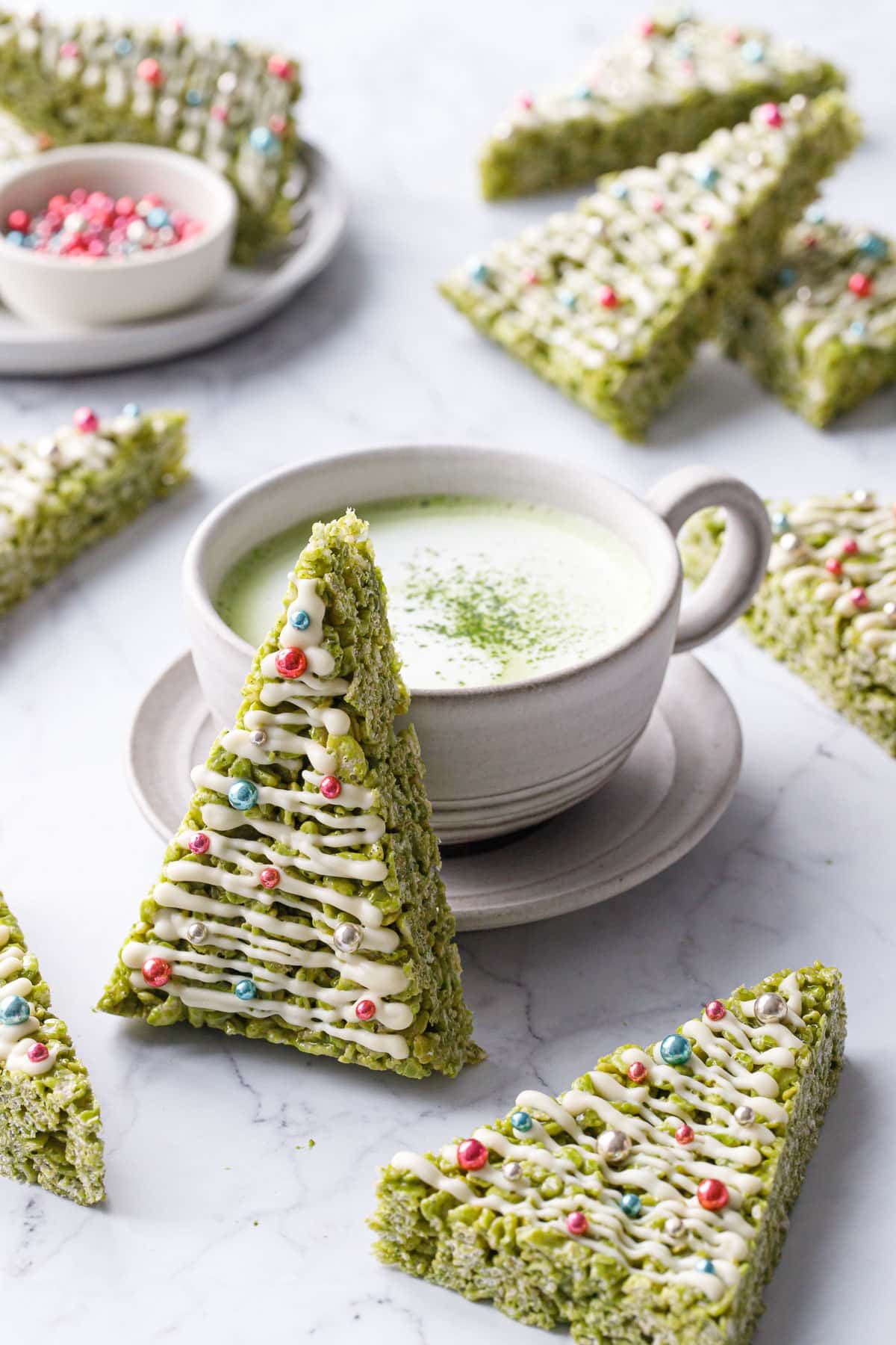 Matcha Rice Crispy Treats cut into triangles and decorated like Christmas trees, one leaning up against a mug of matcha on a marble background.