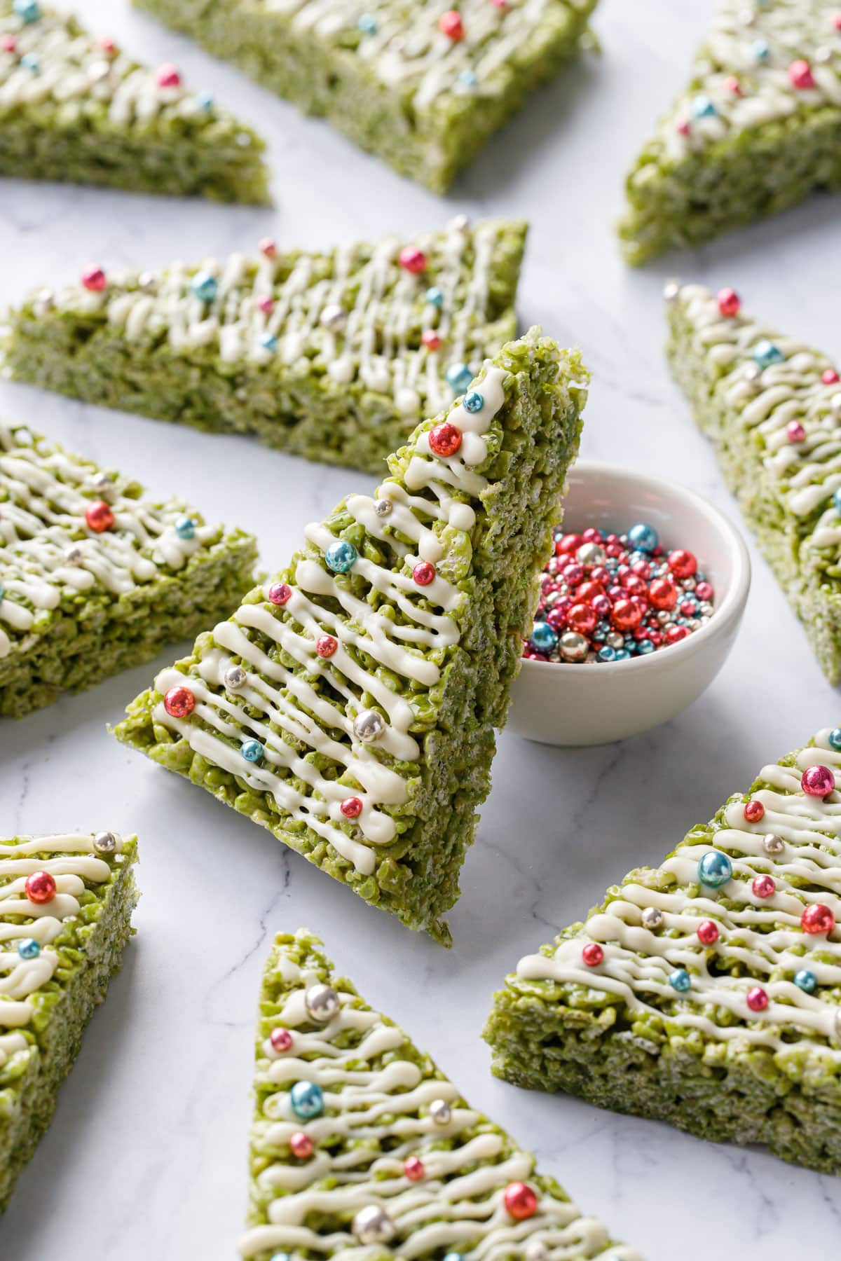 Matcha Rice Crispy Treats cut into triangles and decorated like Christmas trees, one leaning up against a bowl of metallic sprinkles on a marble background.