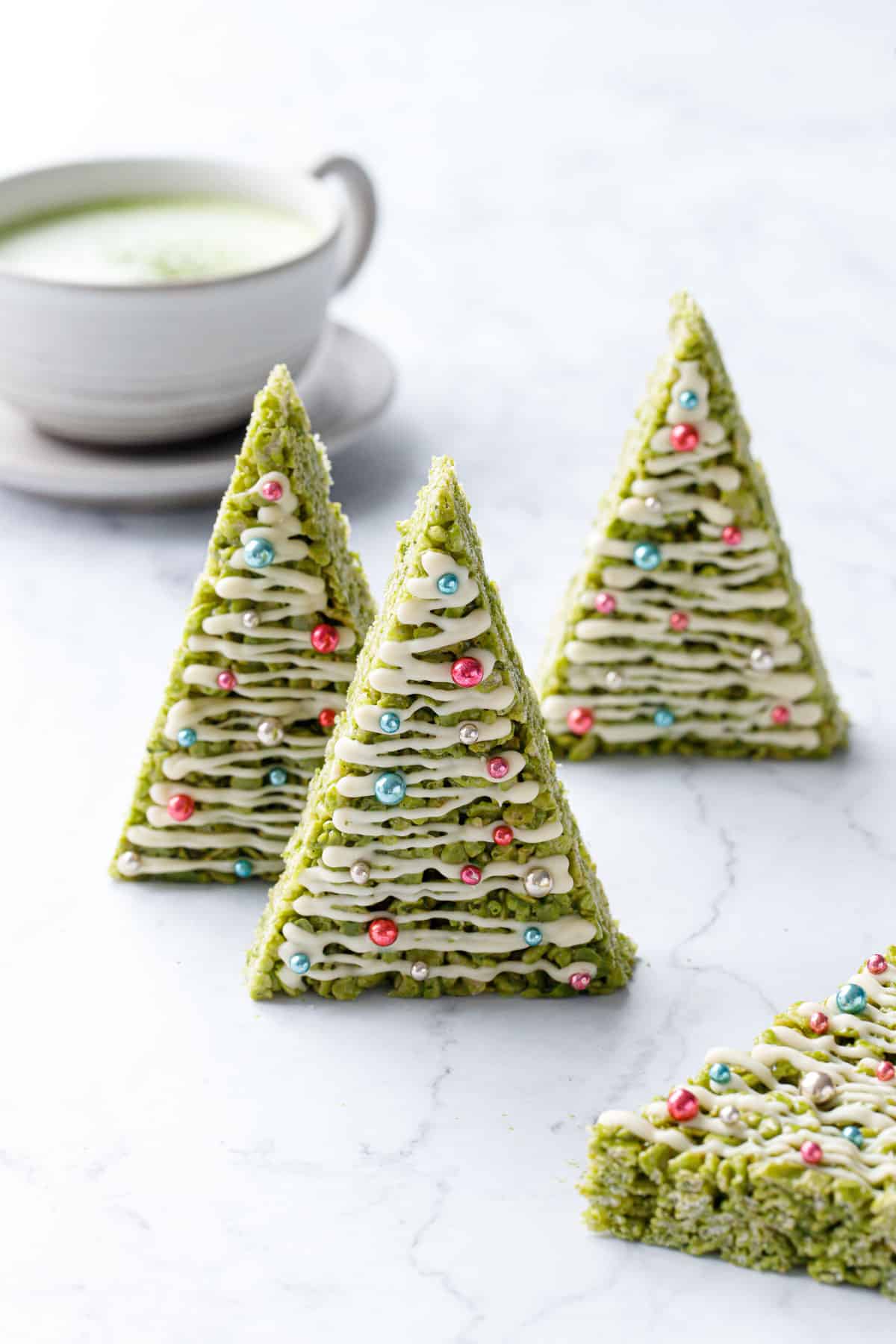 Three Christmas tree shaped Matcha Rice Crispy Treats standing up on a marble background, mug of matcha latte in the background.