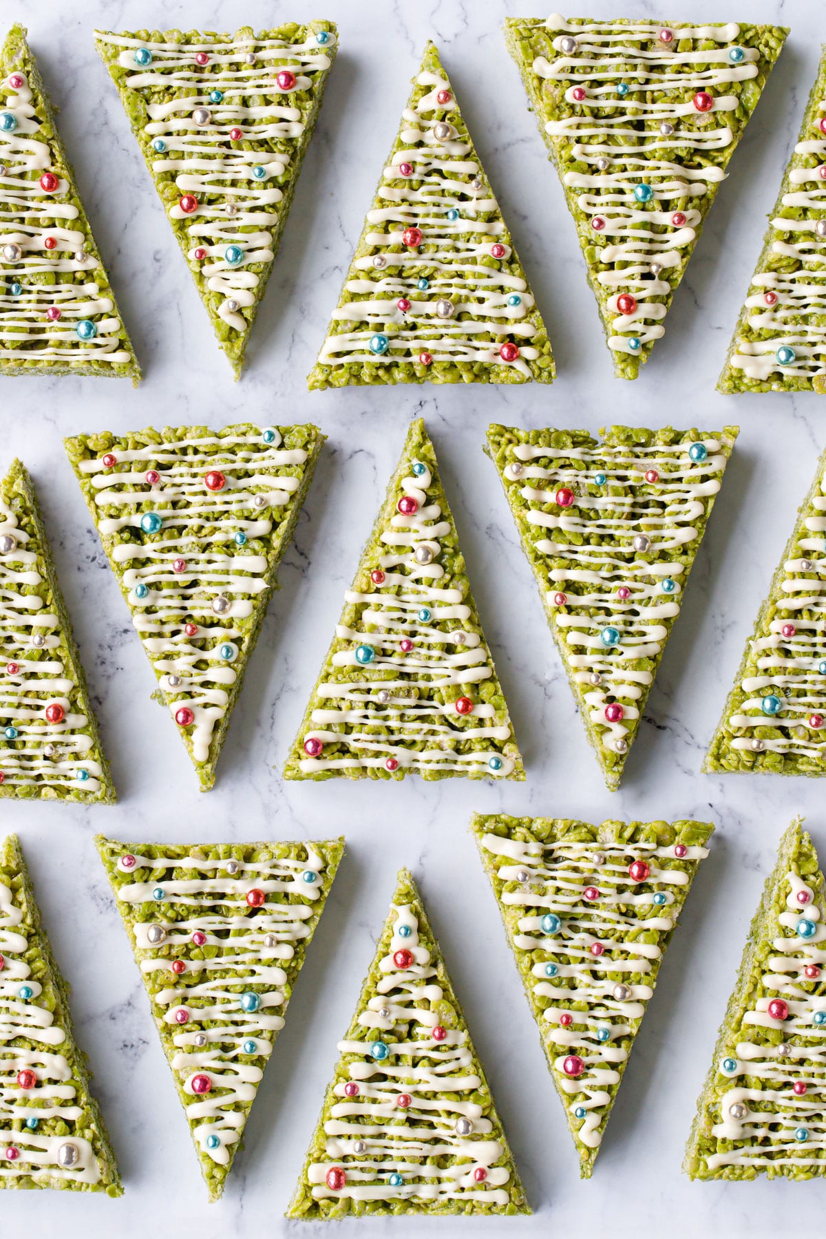 Overhead, rows of triangular Matcha Rice Crispy Treats arranged geometrically, decorated to look like Christmas trees with white chocolate drizzle and shiny ball sprinkles.