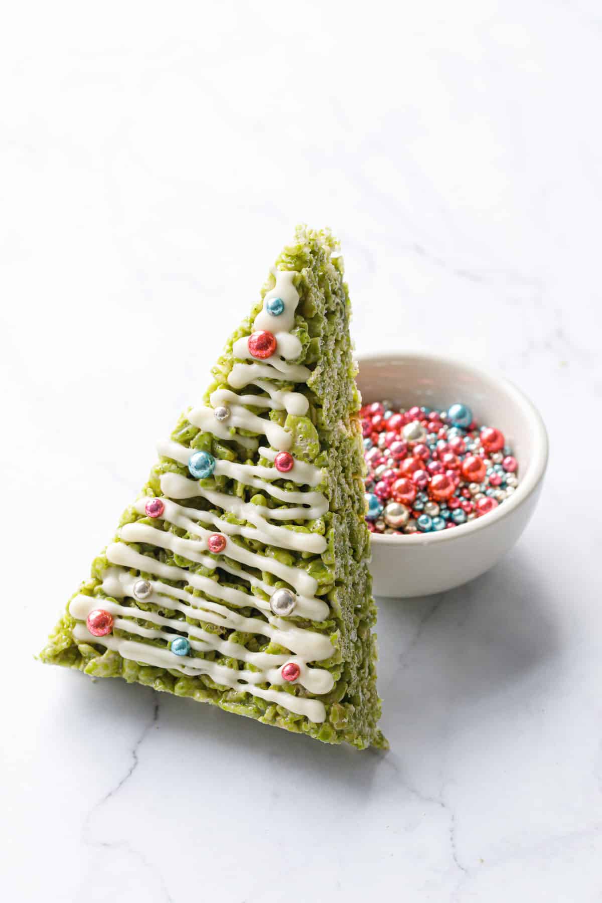Matcha Rice Crispy Treats cut into a triangle and decorated like a Christmas tree, leaning up against a bowl of metallic sprinkles.