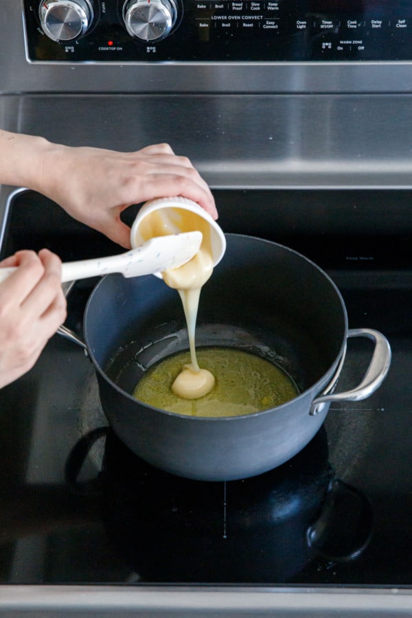 Pouring sweetened condensed milk into a saucepan with melted butter.