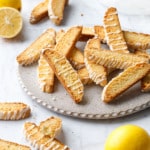 Flat plate with randomly arranged piles of Meyer Lemon Biscotti, with a few lemons (one cut) and a white napkin in the background.