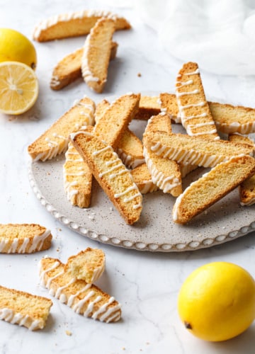 Flat plate with randomly arranged piles of Meyer Lemon Biscotti, with a few lemons (one cut) and a white napkin in the background.