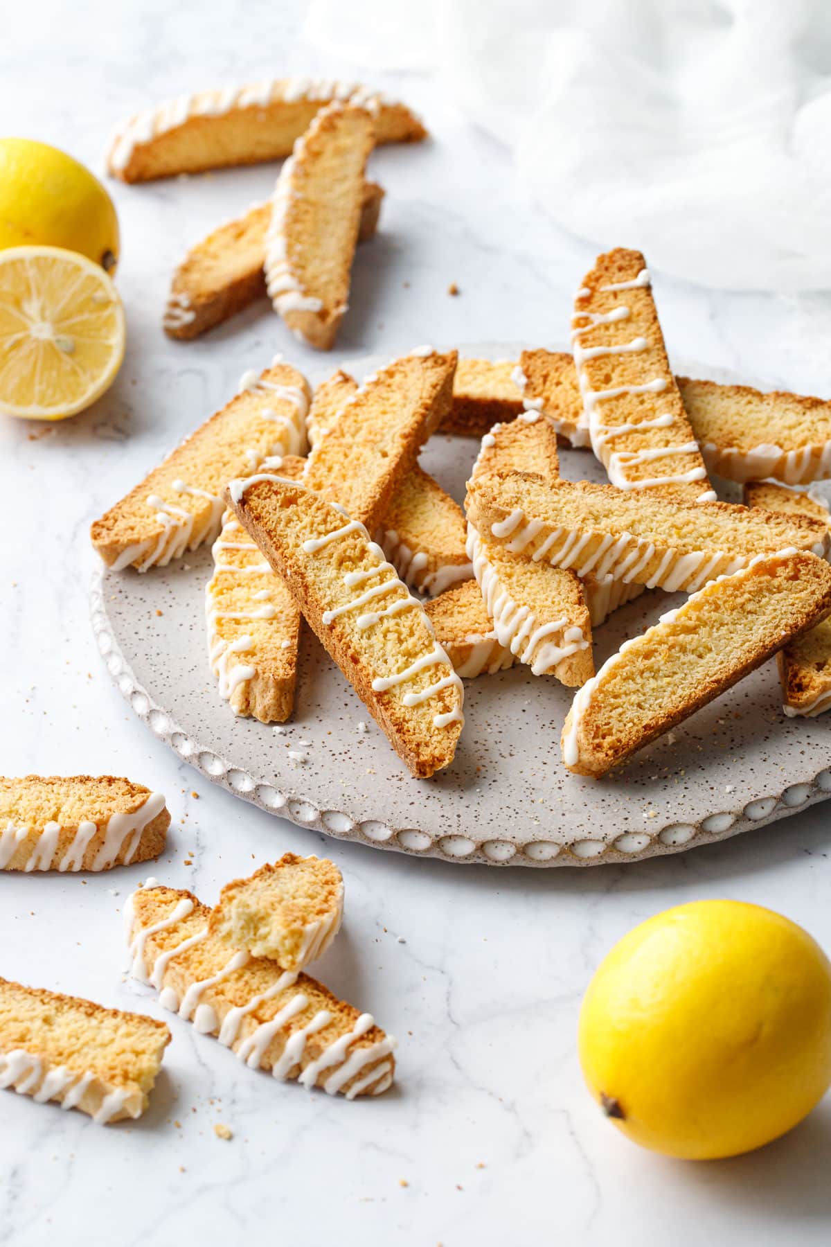 Flat plate with randomly arranged piles of Meyer Lemon Biscotti, with a few lemons (one cut) and a white napkin in the background.