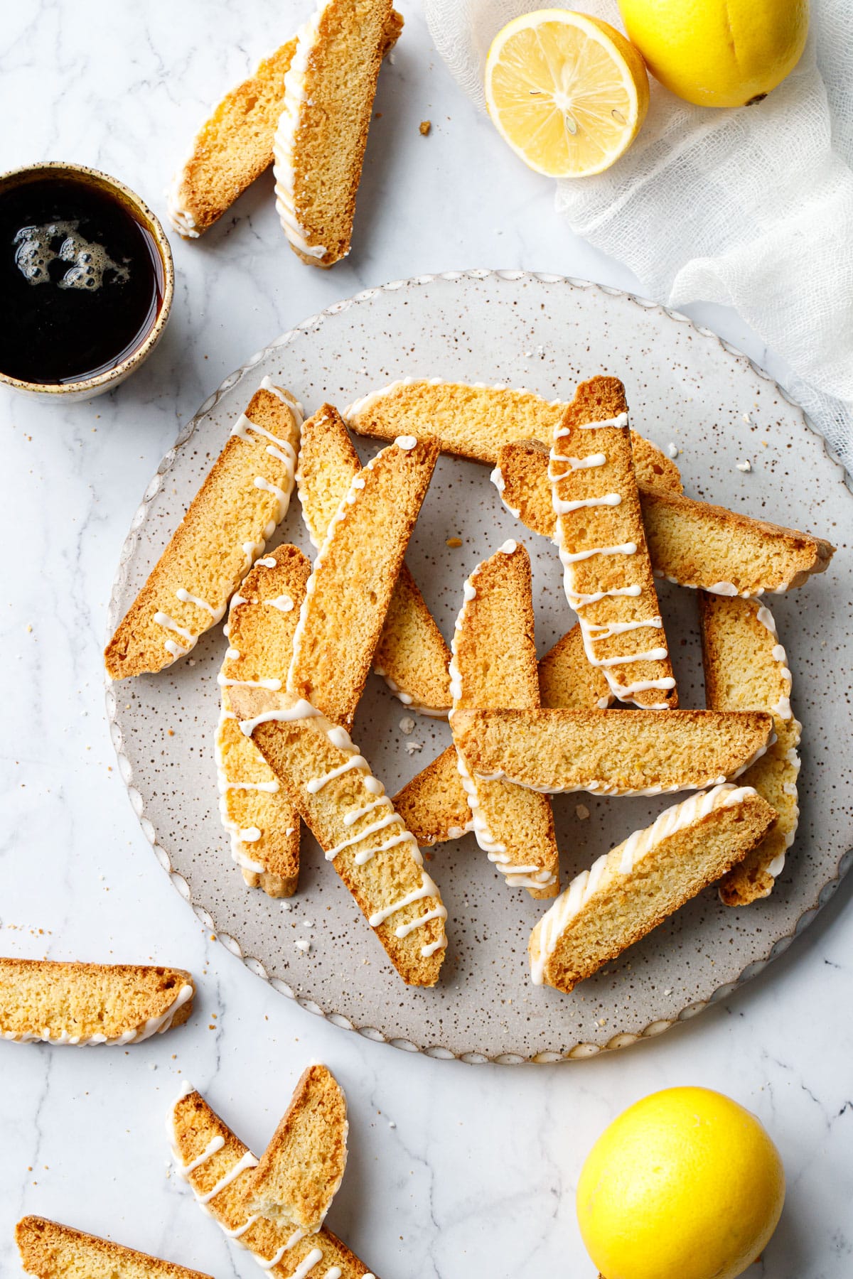 Overhead, round white plate with Meyer Lemon Biscotti piled haphazardly, more cookies scattered around along with a few lemons and a cup of black coffee.