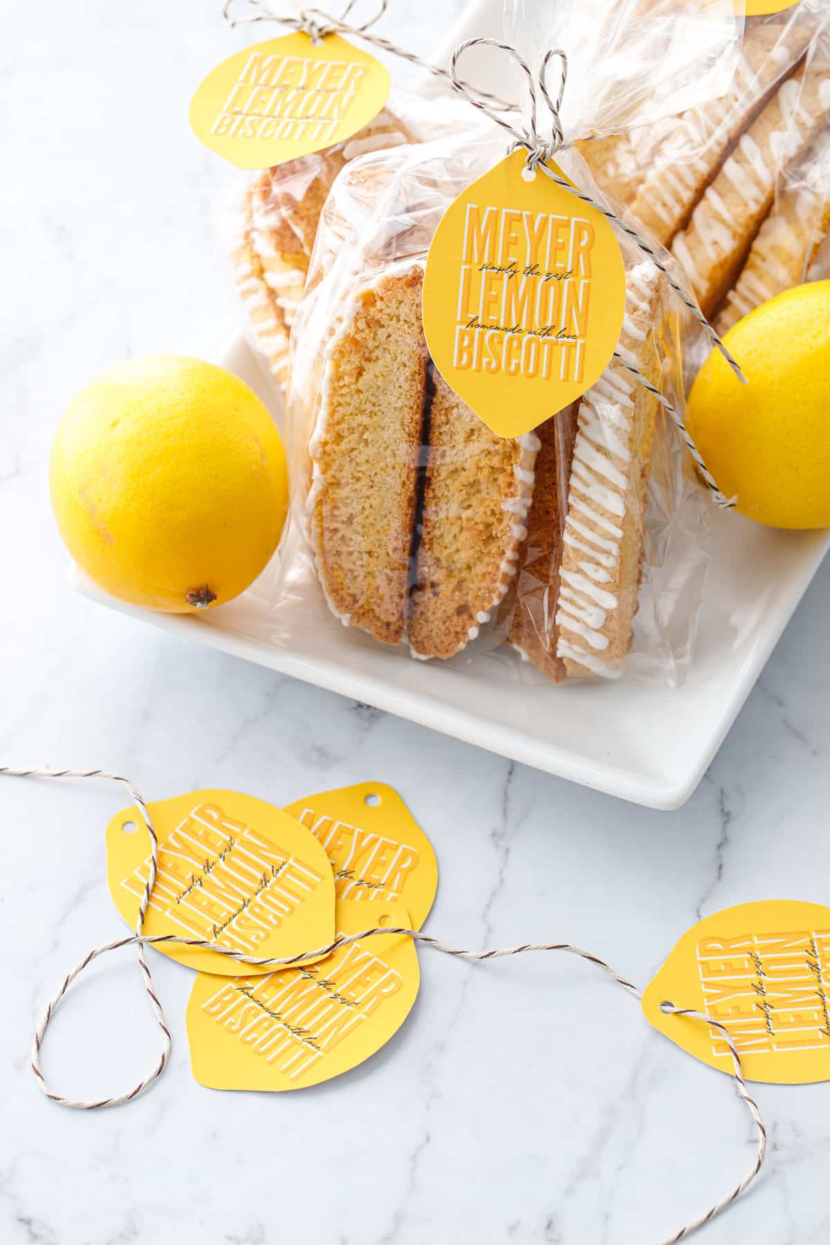 White plate with Meyer Lemon Biscotti in clear cello gift bags, tied with twine and a lemon-shaped gift tag, with a pile of tags in focus in the foreground.