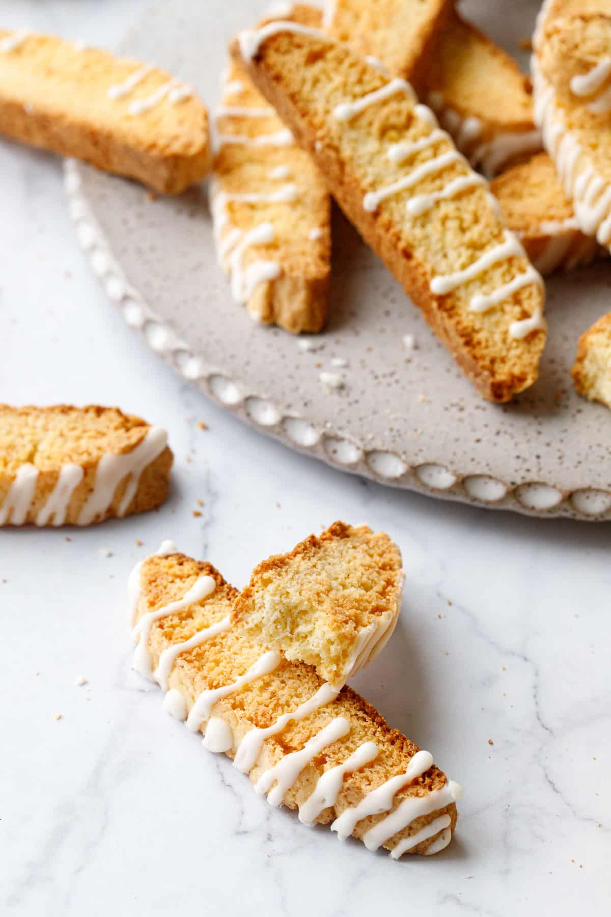 Closeup of a glazed Meyer Lemon Biscotti broken in half to show the interior texture, plate with more biscotti stacked on it in the background.