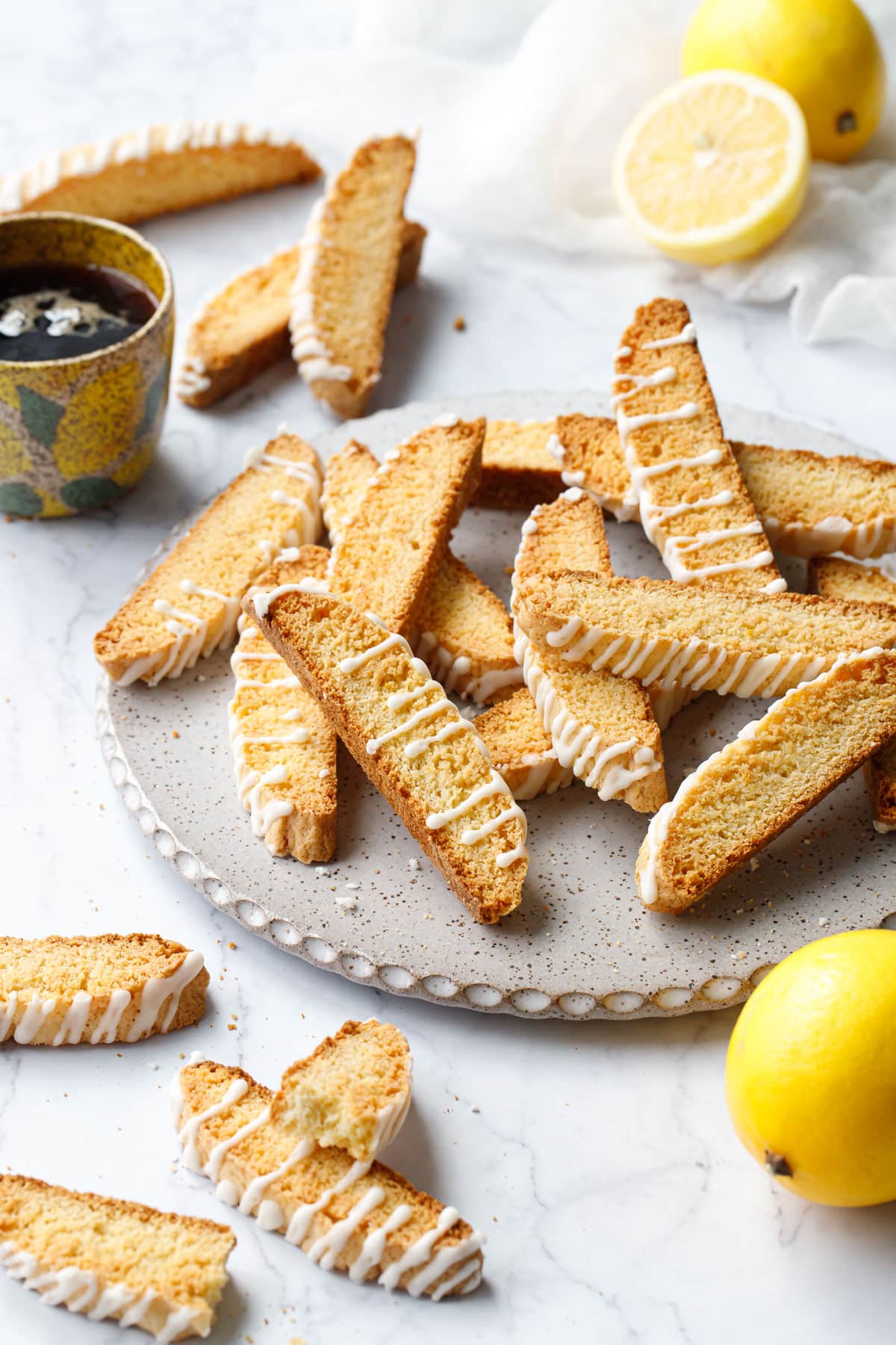 Pile of Glazed Meyer Lemon Biscotti on a ceramic cake plate, with more cookies, a lemon, and a small cup of espresso on the side.