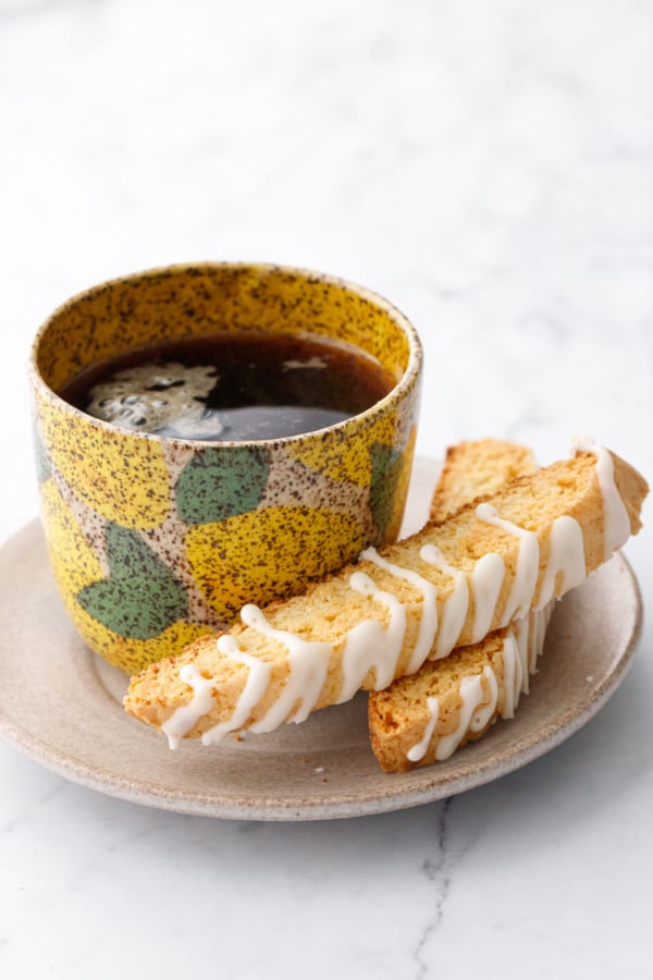 Small plate with a lemon-painted ceramic cup on a saucer with two Meyer Lemon Biscotti cookies drizzled with a white glaze.