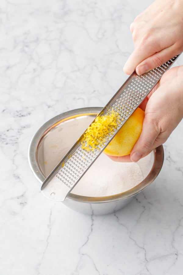 Zesting a lemon with a microplane grater over a bowl with granulated sugar.
