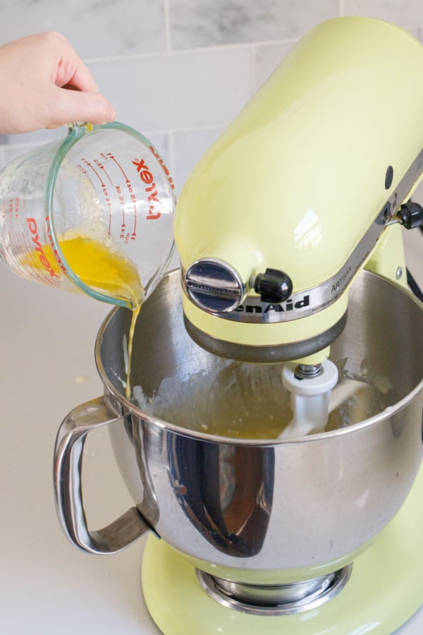 Drizzling melted butter from a glass measuring cup into stand mixer bowl with mixer running on low.