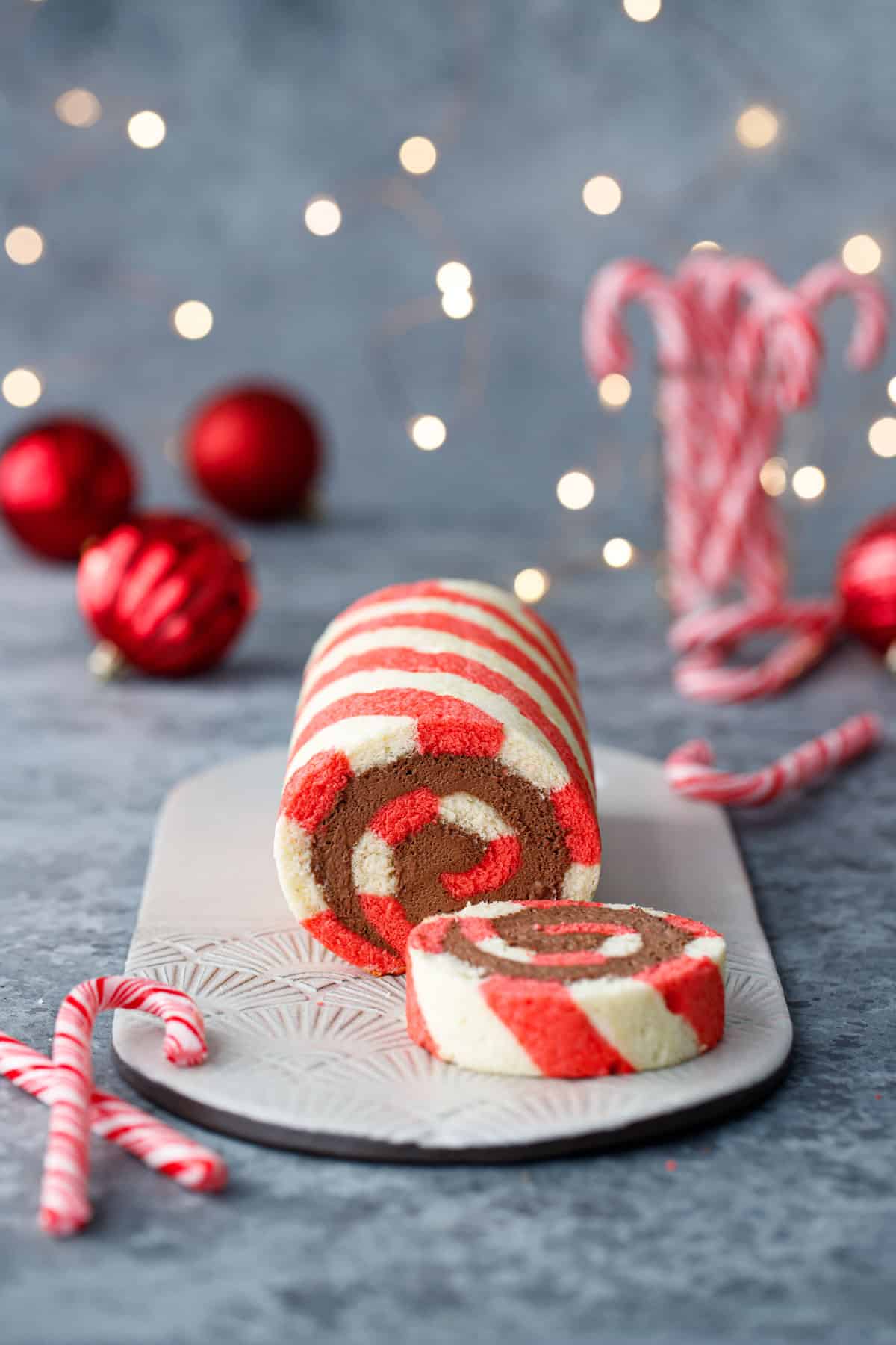 Red and white striped cake roll with one slice cut off to show a perfect spiral of chocolate peppermint whipped cream filling, with candy canes, red ornaments, and Christmas lights out of focus in the background.