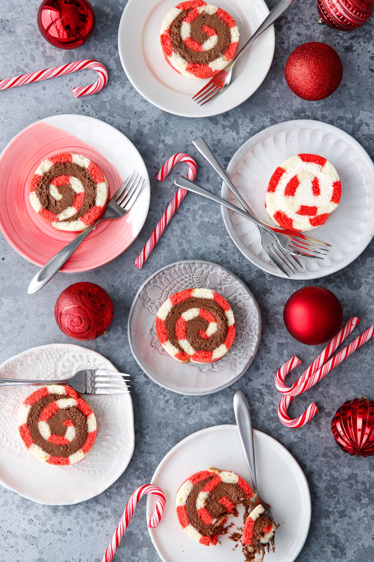Birds-eye view of multiple plates with slices of Peppermint Swiss Cake Rolls with swirls of chocolate and white chocolate whipped cream filling, with some candy canes and red ornaments scattered around.