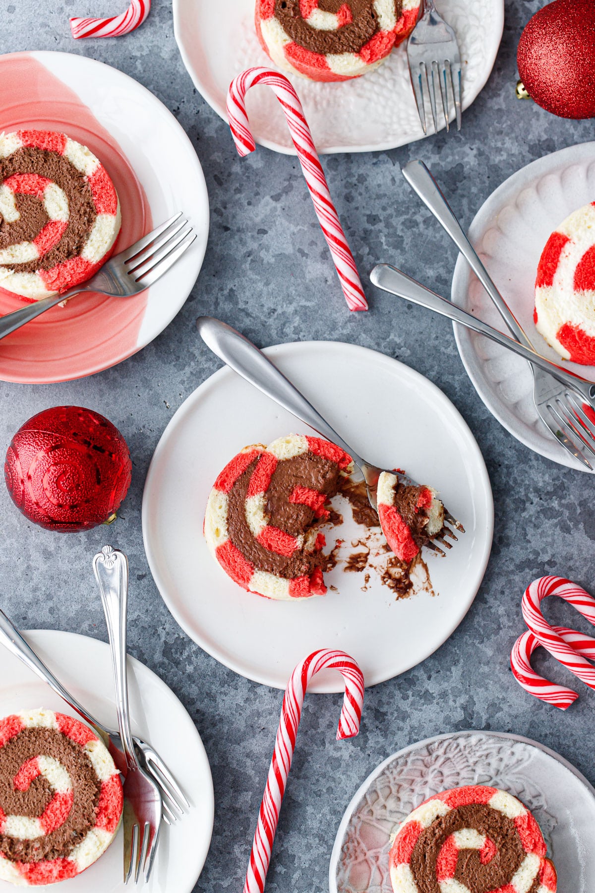 Birds-eye view of multiple plates with slices of Peppermint Swiss Cake Rolls with swirls of chocolate whipped cream filling, one slice smashed a bit with a fork, and a few candy canes and red ornaments scattered around.