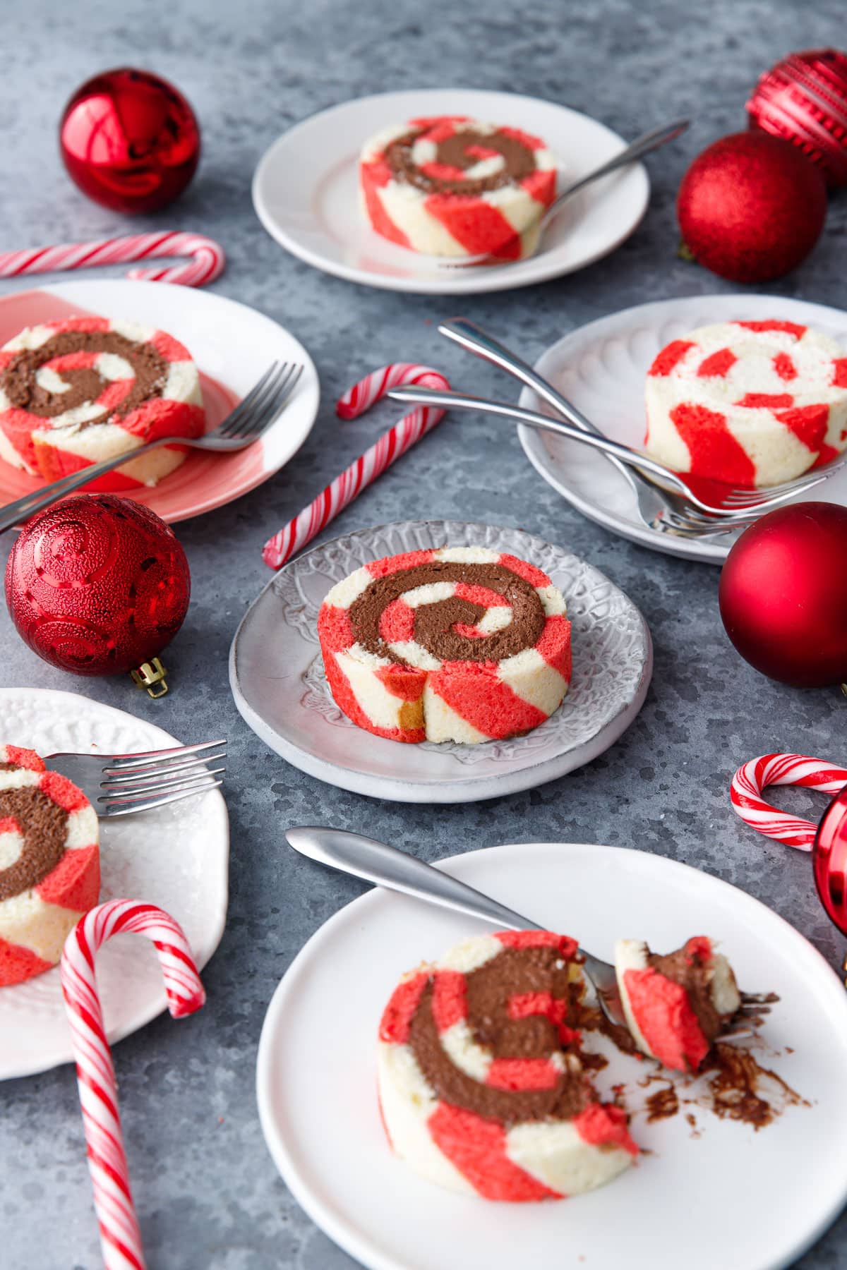 Different styles of ceramic plates with slices of Peppermint Swiss Cake Rolls with both chocolate and white chocolate whipped cream filling, and candy canes and red ornaments scattered around.