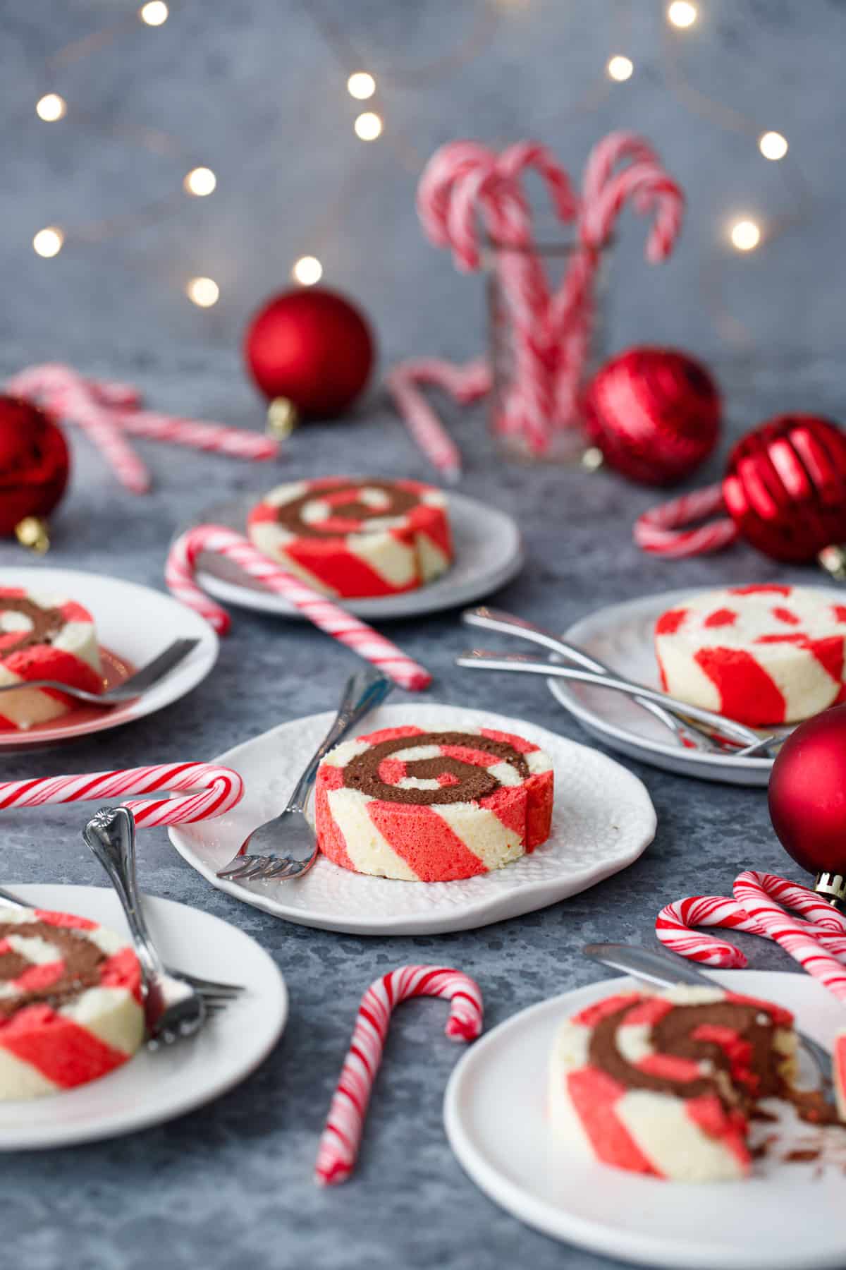 Slices of red and white striped Peppermint Cake Rolls on multiple different plates, with out of focus Christmas lights, candy canes, and red ornaments scattered around.