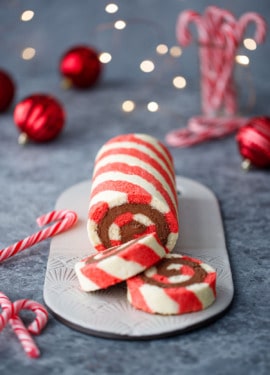 Red and white striped cake roll with chocolate peppermint whipped cream filling, with candy canes, red ornaments, and Christmas lights out of focus in the background.