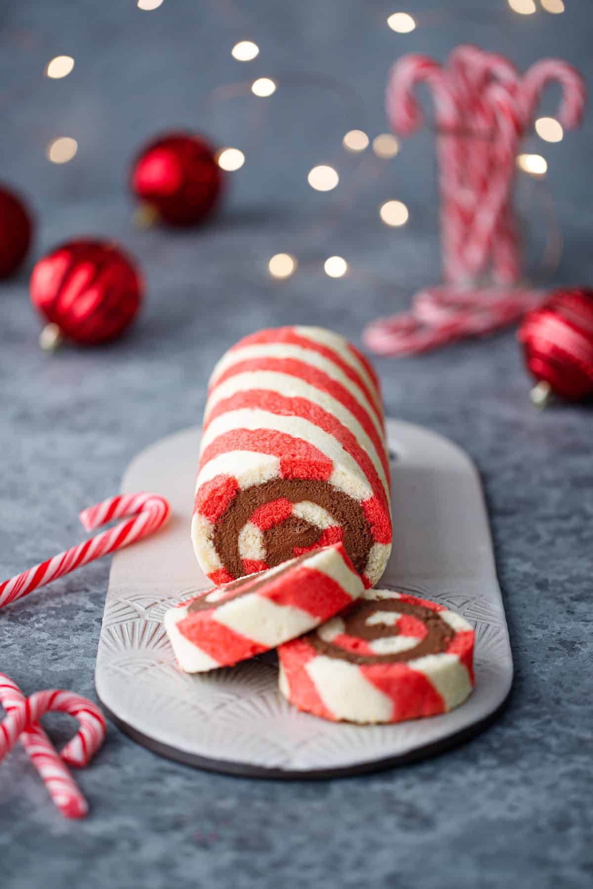 Red and white striped cake roll with chocolate peppermint whipped cream filling, with candy canes, red ornaments, and Christmas lights out of focus in the background.