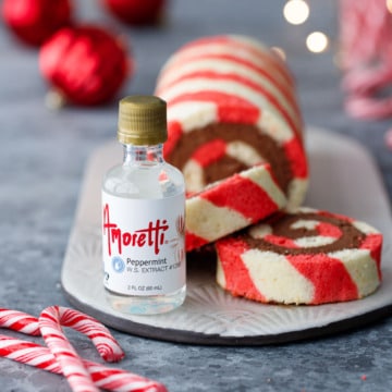 Closeup of Amoretti peppermint extract bottle with Peppermint Striped Cake Roll, plus candy canes, white Christmas lights, and red ornaments out of focus in the background.