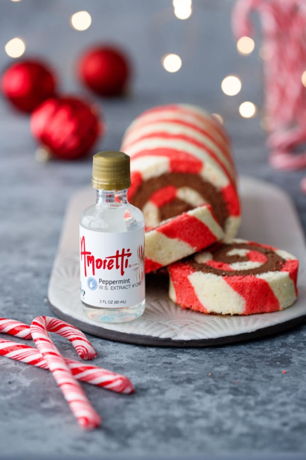 Closeup of Amoretti peppermint extract bottle with Peppermint Striped Cake Roll, plus candy canes, white Christmas lights, and red ornaments out of focus in the background.