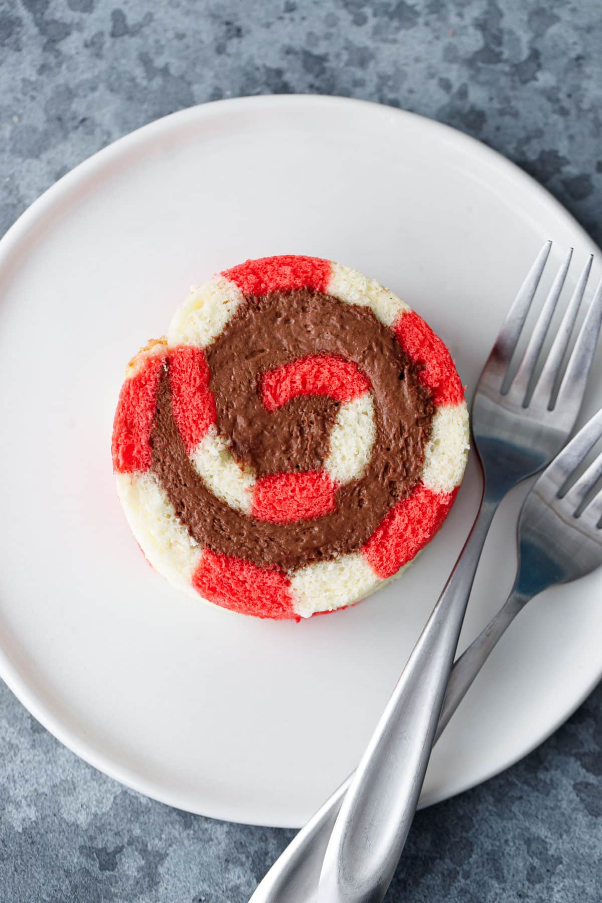 Overhead view of a single slice of peppermint cake roll with a perfect spiral of chocolate whipped cream filling inside on a white plate with two silver forks.